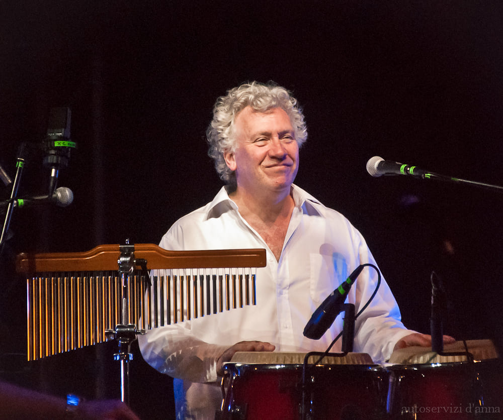 Lächelnder Perkussionist spielt Congas neben einem Windspiel auf der Bühne.