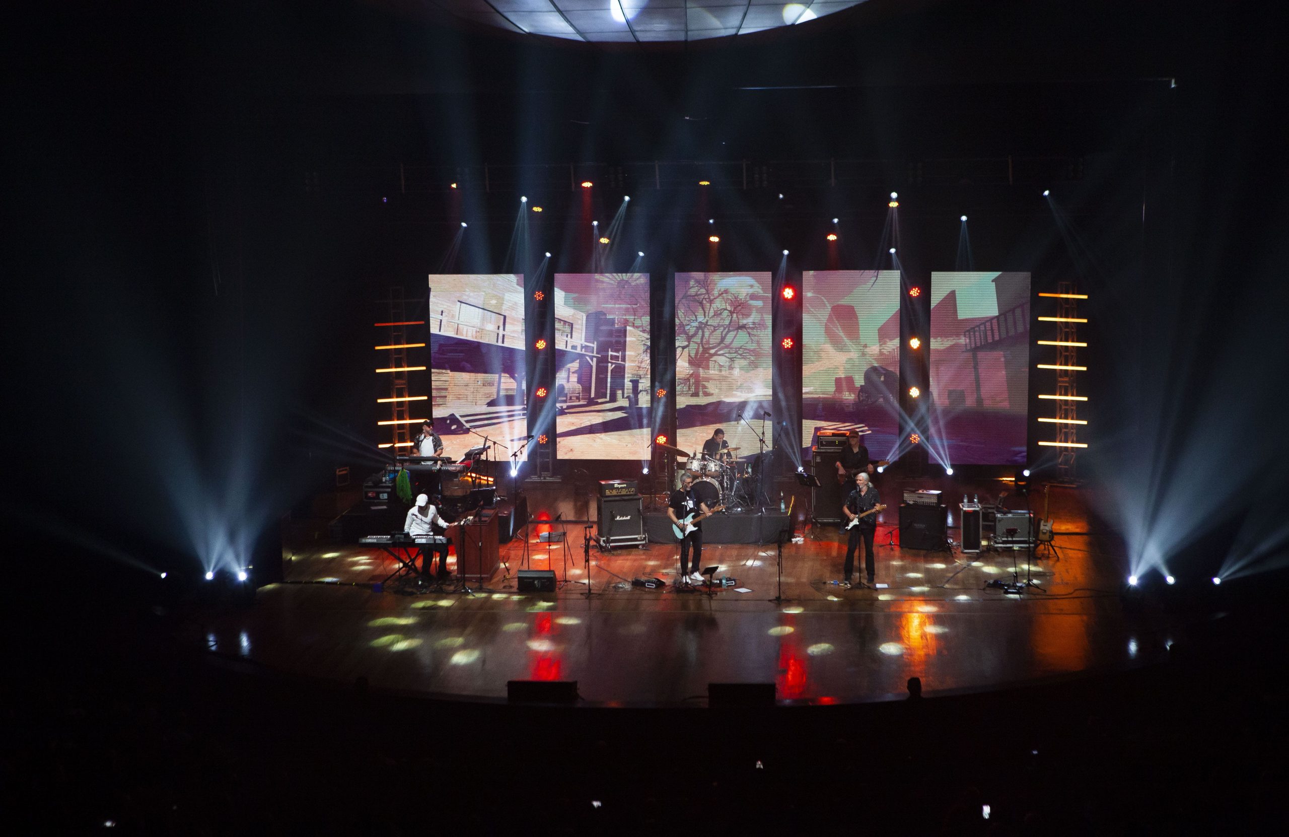 Band Dire Straits Legacy spielt auf einer großen Bühne vor hohen Videowänden und farbigen Scheinwerfern.